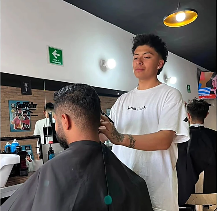 Barbero con playera blanca corta con máquina a un cliente de espaldas, frente al espejo y el muro de ladrillo.
