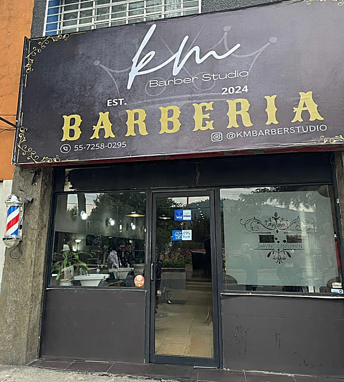 Fachada de KM Barber Studio: lona negra con letras doradas BARBERIA, poste de barbero a la izquierda y ventana con vidrio grabado Shaving Gentlemen.